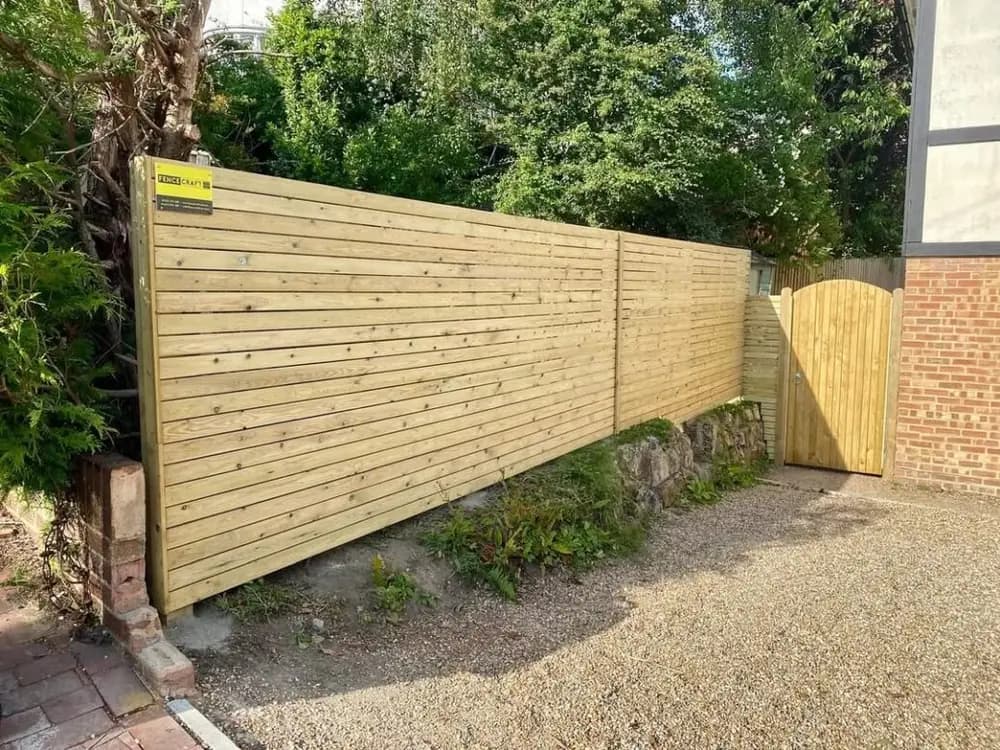 Horizontal slatted fencing with a matching gate beside a gravel path, built by Fencecraft in Kent Horizontal slatted fencing with a matching gate beside a gravel path, built by Fencecraft in Kent